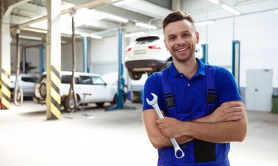 Confident handsome young and experienced car repair worker in work overalls posing against the background of lifted cars in a car service