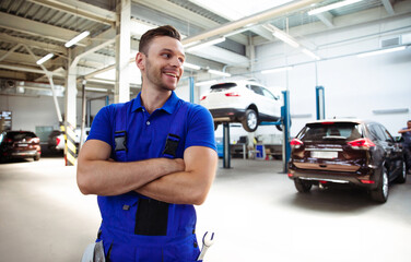 Confident handsome young and experienced car repair worker in work overalls posing against the background of lifted cars in a car service