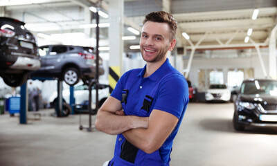 Fototapeta na wymiar Confident handsome young and experienced car repair worker in work overalls posing against the background of lifted cars in a car service