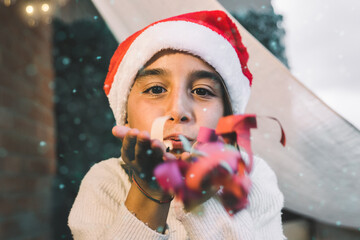 Young girl blowing confetti while having fun during Christmas time - Child wearing Santa Clause hat - Focus on face