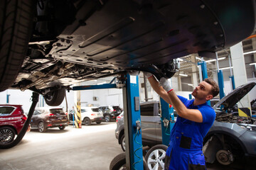 Naklejka premium Cheerful, handsome, and confident car repair specialist in overalls repairs and replaces old parts with new ones in a car on a lift in service