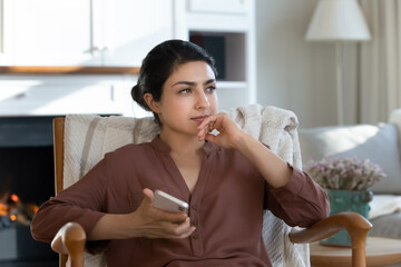 Need to think. Pensive indian lady rest in armchair look aside wait for call choose business entertainment application to download on cell. Meditative young ethnic female hold phone lost in thoughts