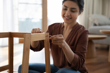 Assembling wood ware. Happy young indian woman flat pack furniture buyer enjoy easy remodeling home making wooden shelf. Biracial lady sit on floor hold allen key mount parts together using hex wrench