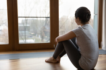 Cozy home. Rear view of dreamy young female in sportswear sitting by large panoramic window hugging knees looking away. Back shot of sporty millennial female relax after fitness training. Copy space
