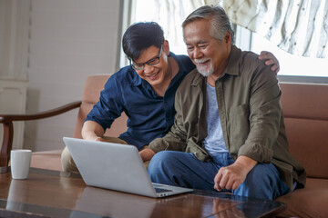 happy asian adult son and senior father sitting on sofa using laptop together at home . young man...