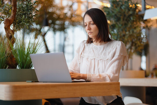 the beautiful and young european woman works on her laptop in a coffee shop or public place. the millennial woman freelancer or finance blogger studies online or meets online. the sad woman works on