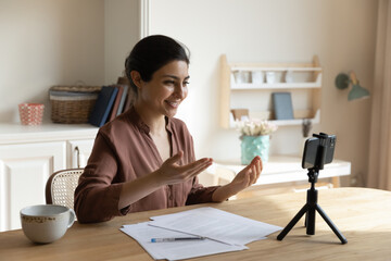 Videoblogging. Happy young lady vlogger of indian ethnicity sit at desk before smartphone on holder...