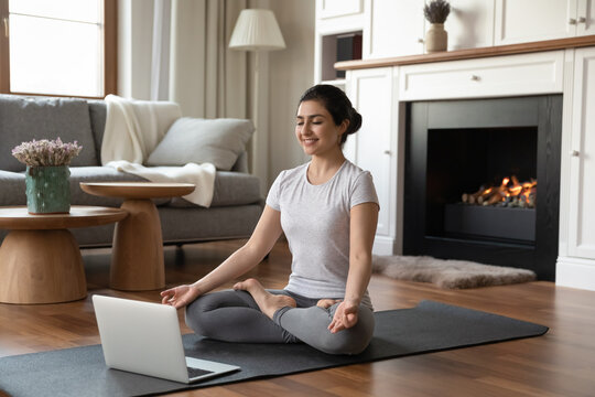 Yoga at home. Young sporty indian female practice sukhasana position meditate on carpet before modern laptop computer at luxury living room. Fit mixed race woman do gymnastic exercises indoor using pc