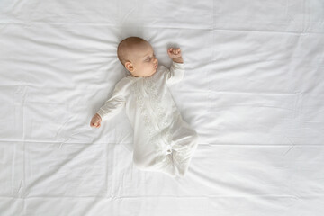 Peaceful adorable baby in bodysuit sleeping on double bed with white sheet. Top view of few month sleepy infant boy or girl lying on back mattress. Child care daily routine concept