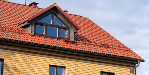 Fototapeta premium Roof of cottage made of ceramic tiles with drains, slopes, snow retainers, tides, chimney, dormer windows against blue sky. Sloping roof with metal coating of residential building with windows