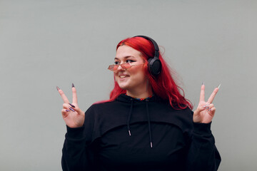European plus size woman in headphones showing V victory fingers sign. Young red pink haired body...