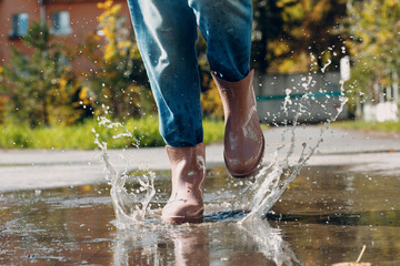 Woman wearing rain rubber boots walking running and jumping into puddle with water splash and drops in autumn rain.