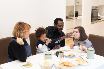 Happy interracial family with young children having breakfast in the kitchen, mixed-race family having fun at the table, moments of tenderness between the parents while the children eat happily