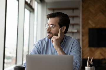 Pensive young Caucasian man distracted from computer job look in window distance thinking planning. Thoughtful male employee work on laptop feel bored unmotivated in office. Boredom concept.