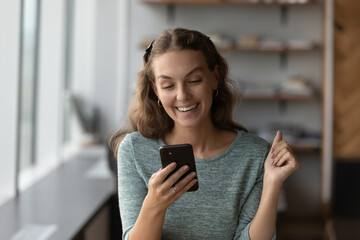 Overjoyed millennial Caucasian woman look at smartphone screen triumph read good news online or get...