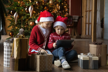 Fototapeta na wymiar Full length happy small children in Santa Claus hats sitting on warm floor near decorated Christmas trees and gifts, involved in playing games together on digital computer tablet, tech addiction.