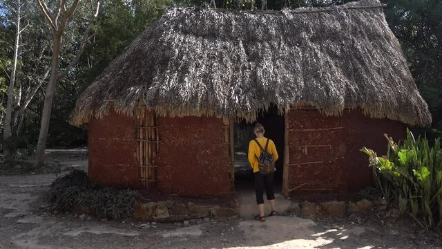 Woman tourist walks into the Chichen Itza traditional pre Columbian Mayan hut home site at historical park.