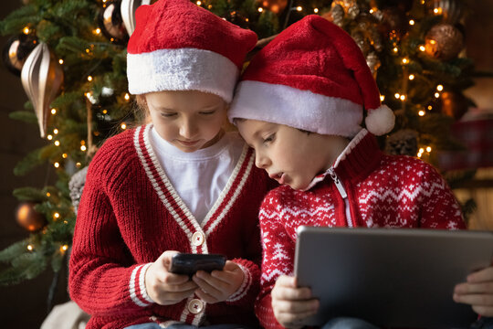 Happy little boy and girl siblings in Santa Claus hats sitting near Christmas tree, using tech presents involved in playing on mobile phone and digital computer tablet together in living room. - Powered by Adobe