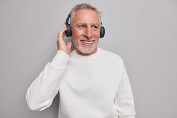 Handsome smiling mature bearded man listens audio track in wireless headphones smiles gladfully...