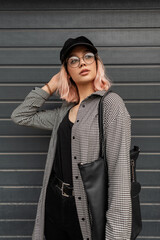 stylish beautiful young woman with glasses and a vintage cap in a fashionable plaid shirt with a bag posing near the gray gate on the street
