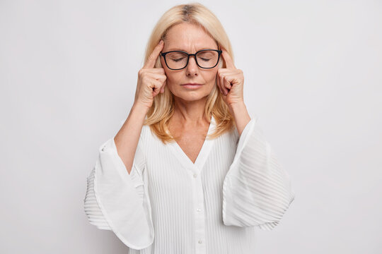 Middle aged blonde attractive woman keeps fingers on temples suffers from headache tries to remember something important stands with closed eyes wears spectacles white blouse faces tough problem