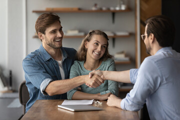 Smiling young Caucasian couple buyers clients shake hand of male real estate agent have meeting in office. Happy millennial man and woman handshake realtor buy rent house, make deal. Rental concept.