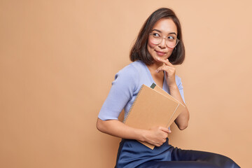 Thoughtful Asian female schoolgirl carries spiral notebooks returns back to school thinks how to improve her knowledge wears spectacles casual clothes sits indoor blank copy space on beige background