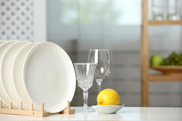 Clean plates in dish drying rack, glasses and lemon on white table indoors - obrazy, fototapety, plakaty