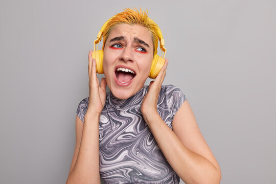 Fashionable punk girl with bright vivid makeup trendy yellow hairstyle sings song enjoys popular music in wireless headphones wears casual t shirt isolated over grey background catches every bit