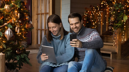 Addicted to tech young couple man and woman using electronic gadgets, congratulating friends online with New Year winter holidays, watching funny Christmas video, relaxing in decorated living room.