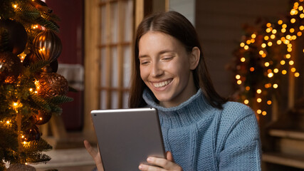 Head shot smiling young caucasian woman in warm winter clothes looking at computer tablet scree, reading Christmas wishes or congratulating friends with New Year night online in social network.
