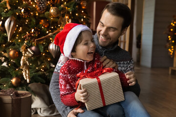 Happy young man embracing little laughing child son, having fun unwrapping gifts sitting on floor near decorated Christmas tree in living room, emotional New Year holiday s family celebration.