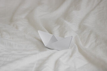 paper origami boat in bed with waves made with bed sheets, natural white crumpled blanket - obrazy, fototapety, plakaty
