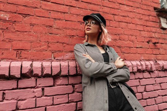 Fashionable beautiful stylish hipster girl with glasses and a cap in casual clothes with a bag stands near a vintage old brick wall