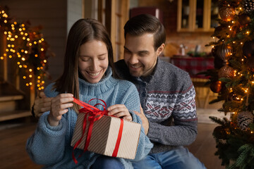 Smiling young man embracing shoulders of happy woman opening wrapped box with New Year Christmas present, feeling excited sitting near decorated tree in living room, winter holidays family celebration