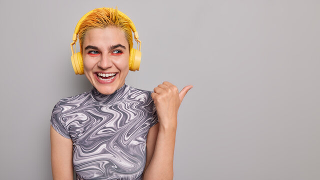 Studio shot of optimistic stylish teenager with trendy hairstyle vivid makeup listens audio podcast indicates thumb away demonstrates copy space for your advertising content shows awesome promo