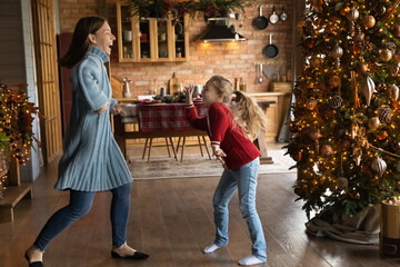 Happy young mother and small laughing daughter wearing warm winter clothes, having fun dancing near...