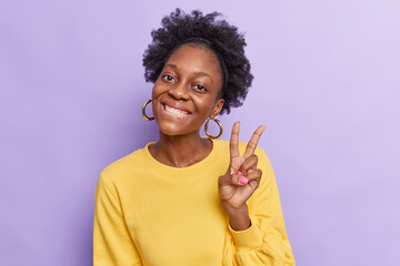 Positive beautiful dark skinned shows peace v sign smiles toothily enjoys nice day gestures victory symbol wears yellow jumper isolated over purple background has good mood carefree expression