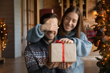 Fototapeta na wymiar Happy young woman covering eyes of smiling curious husband, presenting wrapped Christmas gift, making surprise at New Year night, celebrating winter holidays together near decorated festive tree.
