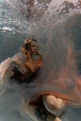 A girl with long dark hair swims underwater in a pink dress and with a crown on her head, like an underwater queen. Fairy tale suitable for advertising	