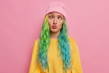 Crazy female student with colored hair has fun alone awkward expression foolishes around after all day ofstudying crosses eyes pouts lips wears hat and jumper poses against pink studio background. - obrazy, fototapety, plakaty