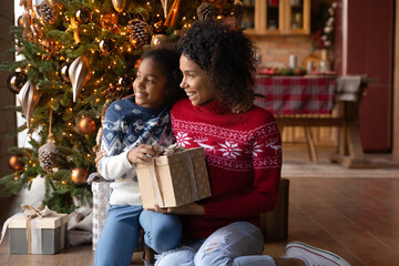 Obraz na płótnie Canvas Happy young African American mom and teen daughter celebrate New Year holidays at home look in distance dreaming. Smiling biracial mother and child enjoy cozy Christmas winter holidays together.