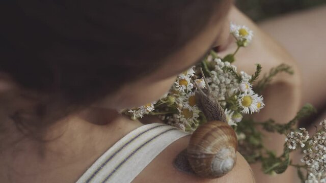 Concept of nature, cosmetology, spa, beauty. Woman holding bouquet of wild flowers or daisies in her hands. Close-up of snail slowly crawling along shoulder of young girl. Snail leaves muscus on skin.