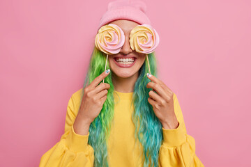 Photo of positive woman has colorful hair covers eyes with caramel candies smiles broadly has sweet tooth enjoys cheat meal day foolishes around wears casual clothes poses against rosy background
