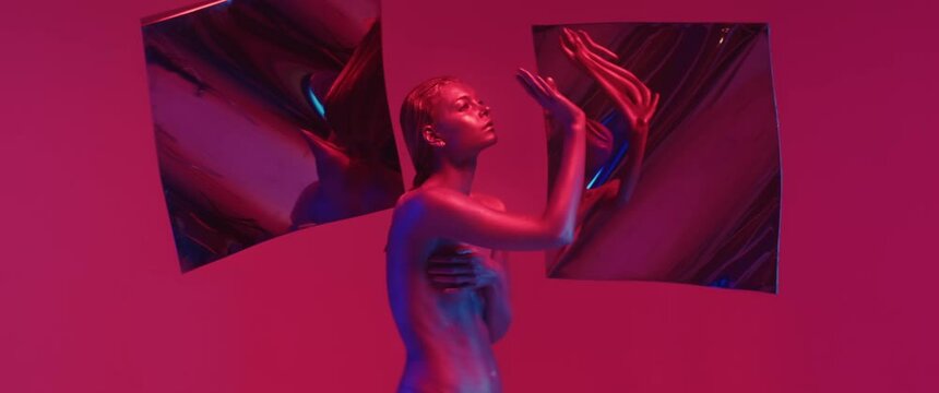 Portrait of attractive girl in colorful neon light, posing sensually and emotionally, making movements with her hands in studio. Female fashion model with face and body covered shiny paint. Anamorphic