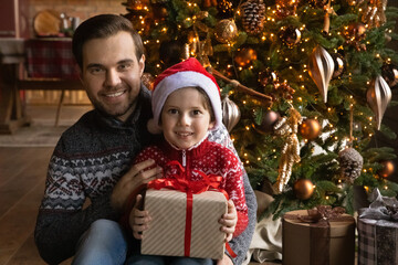Obraz na płótnie Canvas Portrait of happy young father cuddling small excited kid son in Santa Claus hands holding wrapped Christmas gift, sitting together under decorated festive tree on floor, looking at camera.