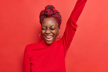 Overjoyed dark skinned young woman keeps arm raised feels upbeat giggles positively wears casual turtleneck and scarf over head poses against red background feels very happy. Emotions concept