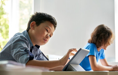 Concentrated diligent teen Asian ethnicity elementary schoolboy studying looking using tablet device sitting in classroom with group of schoolchildren. Modern technologies for education concept.