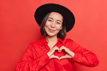 Gentle smiling Asian woman shows heart sign near chest I love you gesture wears black hat and shirt...