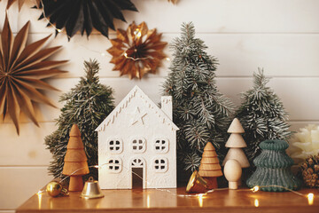 Stylish christmas little house, trees, golden lights on background of paper stars on white wooden...